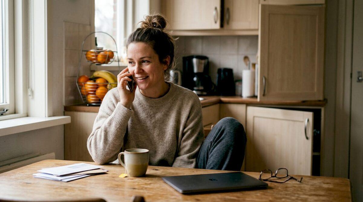 Een vrouw zit thuis aan de keukentafel en is aan het bellen met haar mobiele telefoon.